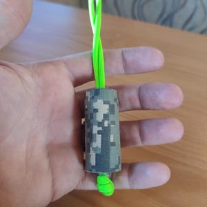 May include: A camouflage-patterned cylinder with a bright green cord attached. The cord is looped and tied at the end.