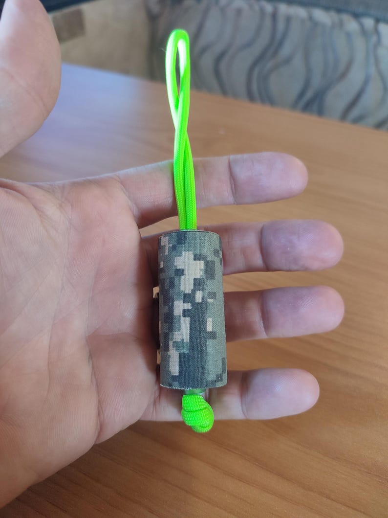 May include: A camouflage-patterned cylinder with a bright green cord attached. The cord is looped and tied at the end.