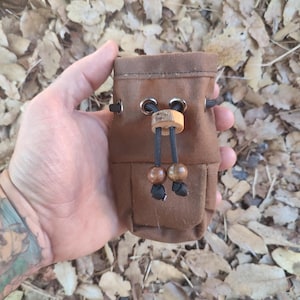 May include: A small, brown suede pouch with a drawstring closure. The pouch has a wooden toggle and decorative wooden beads. The background is a bed of dried leaves.