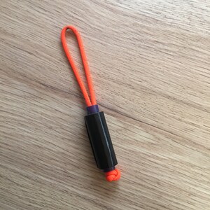 May include: A black and purple cylindrical object with an orange cord loop at the top. The cord is tied in a knot at the bottom of the object.