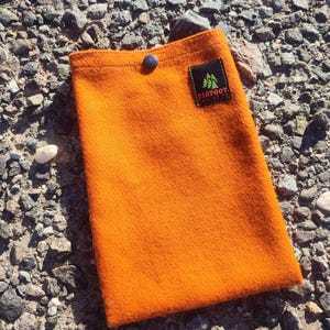 May include: An orange felt sleeve with a black button closure. A small black patch with green trees and the text "BIGFOOT CANVAS CO." is sewn on the front. The sleeve is rectangular and appears to be made of a thick, durable material.