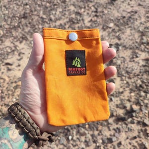 Könnte beinhalten: Ein orangefarbener Canvas-Beutel mit einem weißen Druckknopfverschluss. Ein schwarzes Etikett mit dem Text "BIGFOOT CANVAS CO" und einer Baumgrafik ist auf der Vorderseite aufgenäht. Der Beutel wird in einer Hand gehalten.