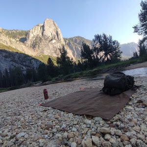 May include: A brown canvas groundsheet laid out on a rocky riverbank in front of a large mountain range. A red water bottle and a black backpack are on the groundsheet.