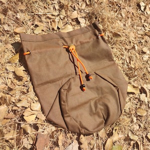 Könnte beinhalten: Eine braune Canvas-Kordeltasche mit orangefarbenen Kordeln und schwarzen Stoppern. Die Tasche liegt auf einem Bett aus trockenen Blättern.