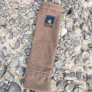 May include: A brown canvas bag with a drawstring closure, resting on a bed of gray and brown rocks. The bag features a rectangular patch with the text "BIGFOOT CANVAS CO." and a green tree logo.