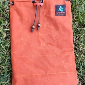 Könnte beinhalten: Eine orangefarbene Canvas-Zugschnur-Tasche mit einer braunen Leder-Zugschnur und einem braunen Leder-Patch mit dem Text "Browns Bushcraft" in Weiß.