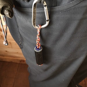May include: A black and blue cylindrical object with a red and black cord attached to a silver carabiner. The carabiner is attached to a black belt loop.