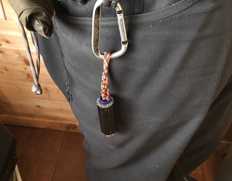 May include: A black and blue cylindrical object with a red and black cord attached to a silver carabiner. The carabiner is attached to a black belt loop.