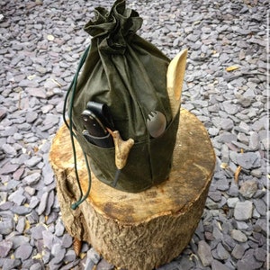 Puede incluir: Una bolsa de lona verde con varios bolsillos, llena de equipo de camping, incluyendo un cuchillo, una cuchara de madera y un tenedor de metal. La bolsa está sentada sobre un tocón de árbol.