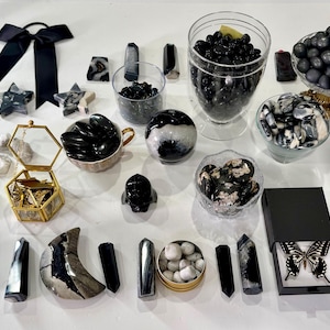 Könnte beinhalten: Eine Sammlung von schwarz-weißen Kristallen, Steinen und Mineralien, arrangiert mit Glasbehältern, einer goldgerahmten Schachtel, einer schwarzen Schleife und einer schwarzen Box mit einem Schmetterlingspräparat. Enthält polierte Kugeln, Türme und Steine.