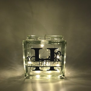 May include: A clear glass block, square-shaped, with the letter "H" and the word "HILL" in black, lit by fairy lights. The block has a decorative design. The lights emit a warm glow, creating a cozy atmosphere.