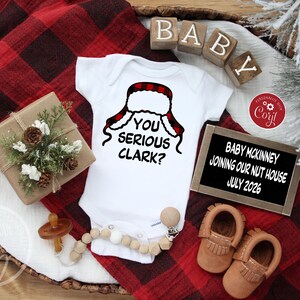 May include: A white baby onesie with the phrase "YOU SERIOUS CLARK?" and a red and black plaid hat design. The image includes baby shoes, a pacifier, and a sign announcing a July 2026 arrival.