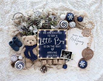 May include: A baby announcement featuring a teddy bear with a blue scarf, blue booties, and decorative ornaments. A sign reads "We are excited to say a Little Boy is on the way! Arriving July 2026." A onesie says "Baby Boy McKinney."