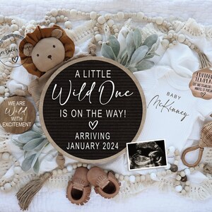 Peut inclure: Un tableau d'affichage noir et blanc avec le texte "A Little Wild One Is On The Way! Arriving January 2024". Le tableau est entour&eacute; d'une couverture blanche, d'un lion en peluche, d'une grenouill&egrave;re pour b&eacute;b&eacute;, d'un hochet en bois et d'une petite pancarte en bois qui dit "We Are Wild With Excitement".