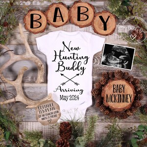 May include: A white onesie with the text "New Hunting Buddy Arriving May 2024" and a pair of crossed arrows. The onesie is surrounded by wooden slices with the letters "BABY" and a wooden slice with the text "BABY MCKINNEY".
