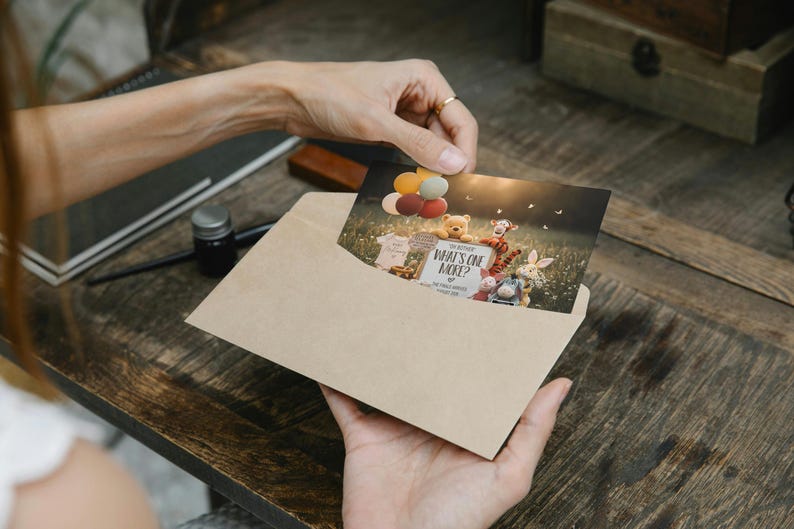 May include: A tan envelope holds a card with Winnie the Pooh characters and the text "What's One More?" Colorful balloons float above the characters. The card is being held by a person.