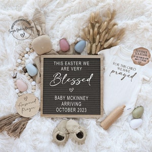 May include: A white onesie with the text "For this child we have prayed" on it. A letter board with the text "This Easter we are very Blessed. Baby McKinney Arriving October 2023". A wooden sign with the text "Answered Prayer".