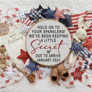 May include: A white baby onesie with the text "BABY McKinney" on it, surrounded by red, white, and blue stars, a wooden rattle, and a sign that reads "Hold on to your sparklers! We've been keeping a little secret. Due to arrive January 2024."