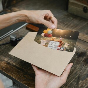 May include: A greeting card featuring Winnie the Pooh characters, balloons, and the text "Our Little Honey is on the way". The card is inside a kraft paper envelope, held by a person. The scene is set in a field with a soft, natural light.