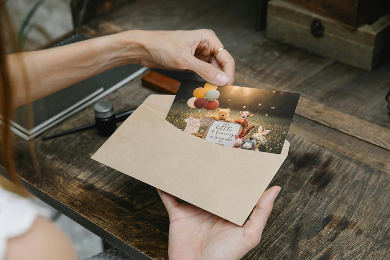 May include: A greeting card featuring Winnie the Pooh characters, balloons, and the text "Our Little Honey is on the way". The card is inside a kraft paper envelope, held by a person. The scene is set in a field with a soft, natural light.