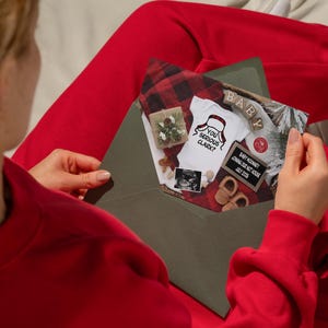May include: An open olive green envelope revealing a pregnancy announcement. Inside, a onesie with the text "You Serious Clark?" is displayed alongside baby shoes, a sonogram, and a small sign.