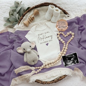 K&ouml;nnte beinhalten: Ein wei&szlig;er Strampler mit dem Text "BABY GIRL McKinney OCTOBER 2023" in schwarzer Schrift mit einem lila Herz. Der Strampler ist umgeben von einer Holzperlenkette, einem kleinen gestrickten Elefanten, einer Holzrassel und einem Paar wei&szlig;er Babyschuhe. Die Gegenst&auml;nde sind auf einer lila Decke mit wei&szlig;en Fransen angeordnet.