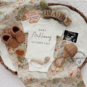 May include: A white baby onesie with the text "Baby McKinney October 2023" on it. The onesie is laid out in a wicker basket with a Winnie the Pooh themed blanket, brown baby shoes, a wooden baby rattle, a pacifier, and a framed ultrasound image.