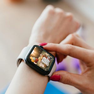 May include: A black smartwatch with a white band is displayed on a wrist. The watch face shows a colorful image with balloons and text. The person's hand with red nail polish is touching the watch.