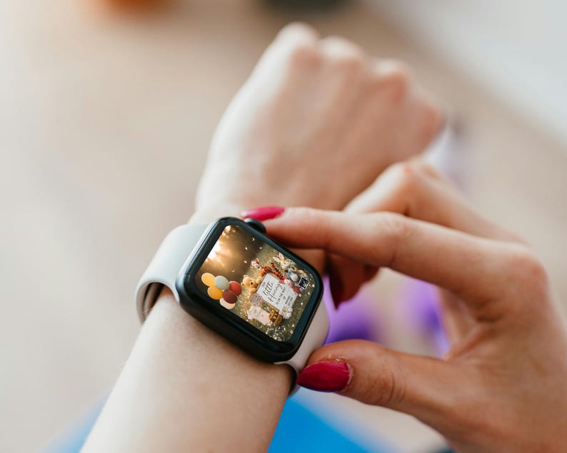 May include: A black smartwatch with a white band is displayed on a wrist. The watch face shows a colorful image with balloons and text. The person's hand with red nail polish is touching the watch.