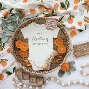May include: A white baby onesie with the text "BABY McKinney NOVEMBER 2023" on it, surrounded by oranges, a wooden rattle, and wooden blocks that spell out "BABY".