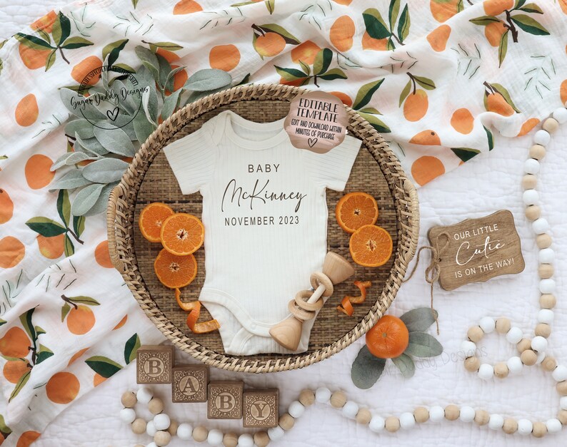 May include: A white baby onesie with the text "BABY McKinney NOVEMBER 2023" on it, surrounded by oranges, a wooden rattle, and wooden blocks that spell out "BABY".