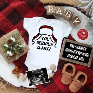 May include: A white baby onesie with a red and black plaid hat design and the text "YOU SERIOUS CLARK?". The image includes a gift box, baby shoes, ultrasound image, and a sign that reads "BABY MCKINNEY JOINING OUR NUT HOUSE DECEMBER 2026".