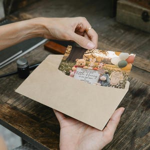 May include: A Winnie the Pooh themed baby announcement card is held in a kraft paper envelope. The card features illustrations of Pooh, Piglet, Tigger, Rabbit, and Eeyore. The card includes the text "We are excited to say our little Honey..." and the baby's arrival date.