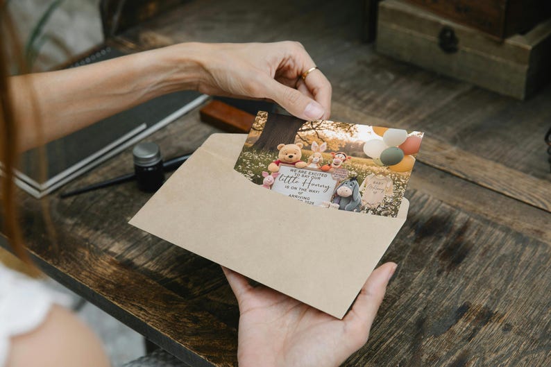 May include: A Winnie the Pooh themed baby announcement card is held in a kraft paper envelope. The card features illustrations of Pooh, Piglet, Tigger, Rabbit, and Eeyore. The card includes the text "We are excited to say our little Honey..." and the baby's arrival date.