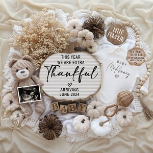 May include: A white baby onesie with the text "Baby McKinney" and two hearts. The onesie is surrounded by fall-themed decorations, including a wooden sign that reads "This year we are extra thankful", a small teddy bear, a white pumpkin, and a wooden sign that reads "Hello Pumpkin".