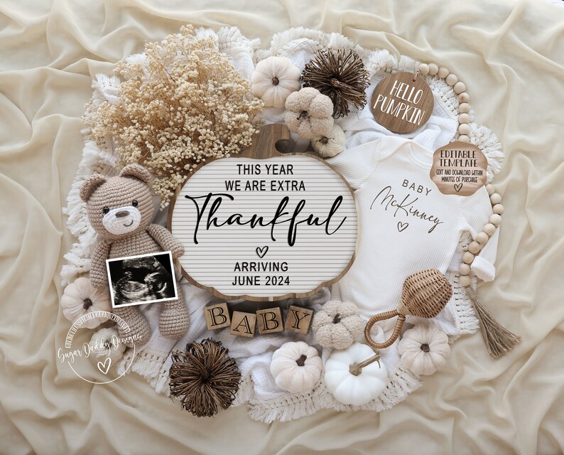 May include: A white baby onesie with the text "Baby McKinney" and two hearts. The onesie is surrounded by fall-themed decorations, including a wooden sign that reads "This year we are extra thankful", a small teddy bear, a white pumpkin, and a wooden sign that reads "Hello Pumpkin".