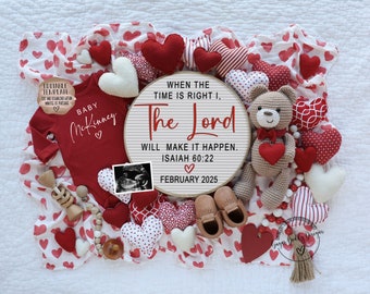 Pode incluir: Um macacão vermelho para bebê com o texto "Baby McKinney" e um coração. Uma placa redonda de madeira com o texto "When the time is right, the Lord will make it happen. Isaiah 60:22. February 2025". A placa é cercada por corações vermelhos e brancos, um urso de pelúcia de crochê e um par de sapatos marrons para bebê.