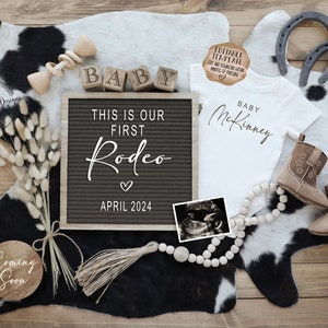 May include: A black letter board with the text "This is our first Rodeo" and the date "April 2024". A white onesie with the text "Baby McKinney" is next to the letter board. A wooden sign with the text "Coming Soon" is in the lower left corner. A cowhide rug is in the background.