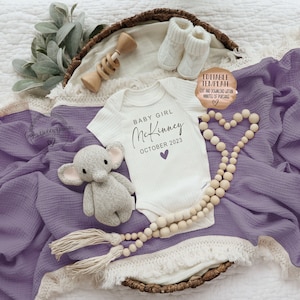 K&ouml;nnte beinhalten: Ein wei&szlig;er Strampler mit dem Text "BABY GIRL McKinney OCTOBER 2023" in schwarzer Schrift mit einem kleinen lila Herz. Der Strampler liegt auf einer lila Decke mit einer Holzperlenkette und einem kleinen gestrickten Elefantenspielzeug.