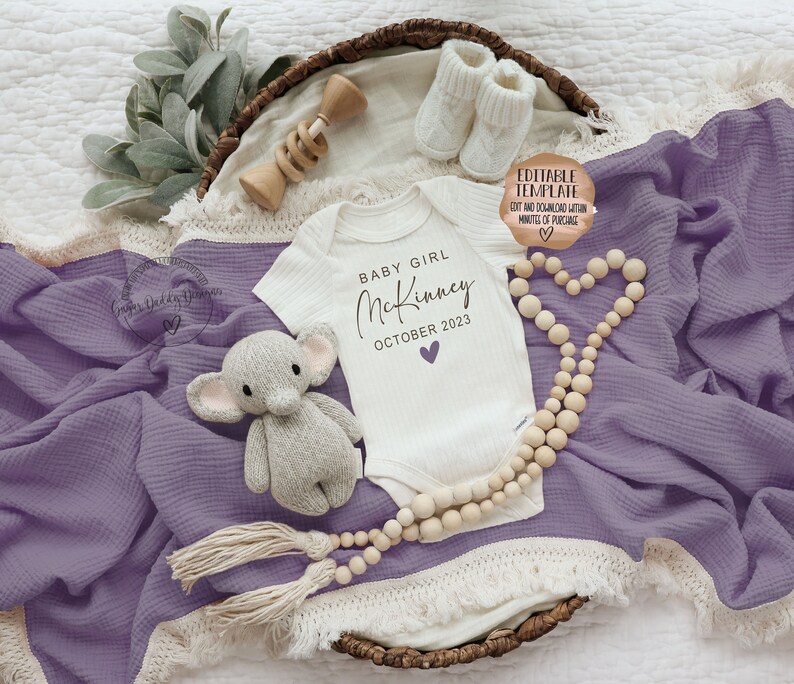 K&ouml;nnte beinhalten: Ein wei&szlig;er Strampler mit dem Text "BABY GIRL McKinney OCTOBER 2023" in schwarzer Schrift mit einem kleinen lila Herz. Der Strampler liegt auf einer lila Decke mit einer Holzperlenkette und einem kleinen gestrickten Elefantenspielzeug.