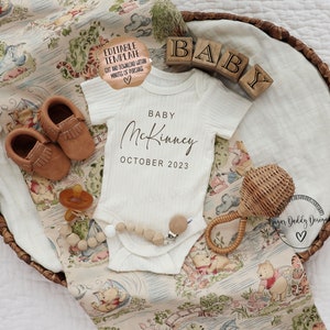 May include: A white baby onesie with the text "Baby McKinney October 2023" in black script font. The onesie is laid out on a Winnie the Pooh themed blanket in a brown wicker basket. There are brown leather baby shoes, a wooden baby rattle, and a pacifier in the basket.