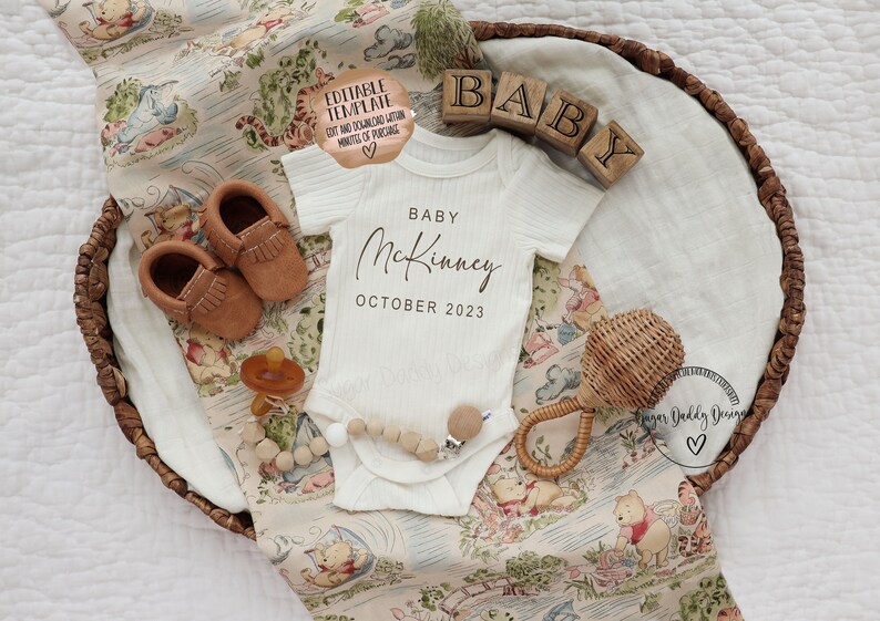 May include: A white baby onesie with the text "Baby McKinney October 2023" in black script font. The onesie is laid out on a Winnie the Pooh themed blanket in a brown wicker basket. There are brown leather baby shoes, a wooden baby rattle, and a pacifier in the basket.