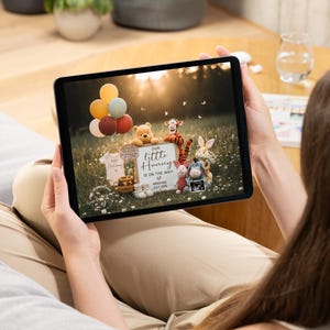 May include: A tablet displays a pregnancy announcement with Winnie the Pooh characters. The announcement includes a sign that reads "Our Little Hunny is on the way!" with the expected arrival date of July 2025. Balloons and baby clothes are also visible.