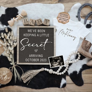 May include: A black letter board with the text "We've been keeping a little secret arriving October 2023".  The letter board is surrounded by a variety of baby-themed items, including a white onesie with the text "Baby McKinney", a pair of brown cowboy boots, a wooden sign with the text "This is our first rodeo", and a string of wooden beads.