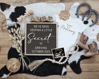 May include: A black letter board with the text "We've been keeping a little secret arriving October 2023".  The letter board is surrounded by a variety of baby-themed items, including a white onesie with the text "Baby McKinney", a pair of brown cowboy boots, a wooden sign with the text "This is our first rodeo", and a string of wooden beads.