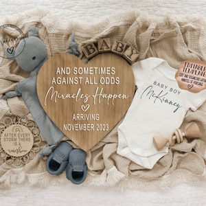 May include: A wooden heart-shaped sign with the text "And sometimes against all odds miracles happen" and "Arriving November 2023". A white onesie with the text "Baby Boy McKinney" is next to the sign. A wooden toy and a blue baby shoe are also in the image.