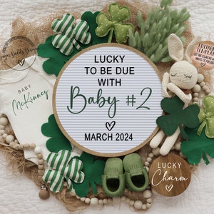 Puede incluir: Un body blanco con el texto "BABY McKinney" en &eacute;l, un letrero de madera con el texto "LUCKY TO BE DUE WITH Baby #2 MARCH 2024", un conejito de crochet y un letrero de madera con el texto "LUCKY Charm" rodeado de tr&eacute;boles verdes.