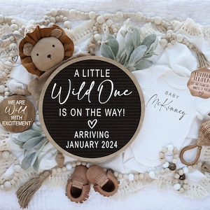 Peut inclure: Un tableau d'affichage noir et blanc avec le texte "A Little Wild One Is On The Way! Arriving January 2024". Une grenouill&egrave;re blanche avec le texte "Baby McKinney" est &agrave; c&ocirc;t&eacute; du tableau d'affichage. Un lion en peluche, un petit hochet en bois et une guirlande de perles en bois sont &eacute;galement pr&eacute;sents sur l'image.