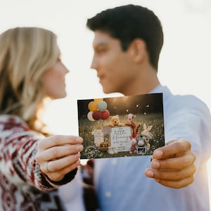 May include: A couple holds a card announcing a pregnancy. The card features Winnie the Pooh characters, balloons, and the text "Our Little Hunny is on the way!" The background is a sunny outdoor scene.