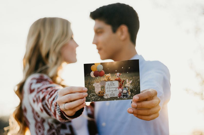 May include: A couple holds a card announcing a pregnancy. The card features Winnie the Pooh characters, balloons, and the text "Our Little Hunny is on the way!" The background is a sunny outdoor scene.